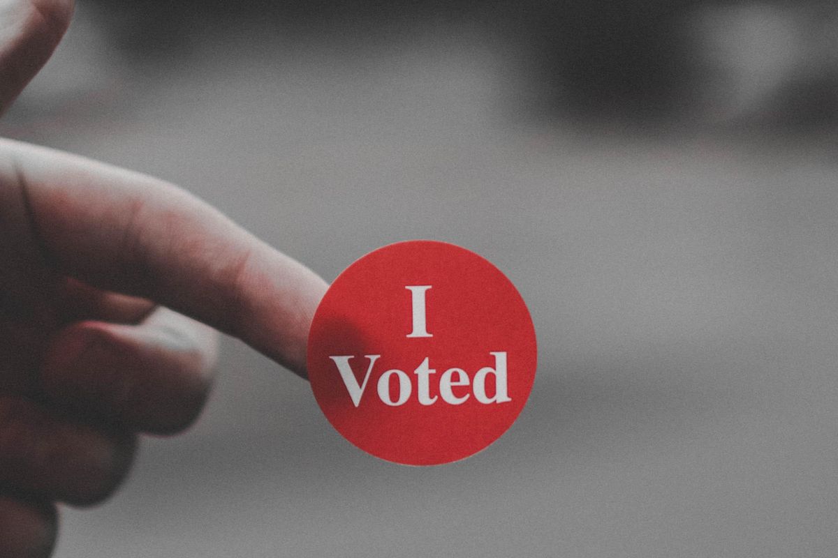 Close up picture of a red circular sticker with white writing that says "I voted" stuck to a finger.
