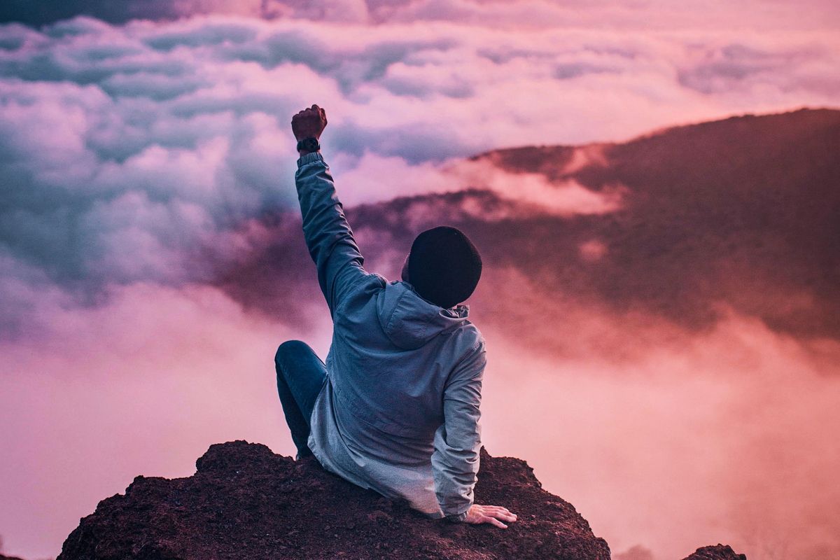 man on mountain with left fist in the air towards the clouds