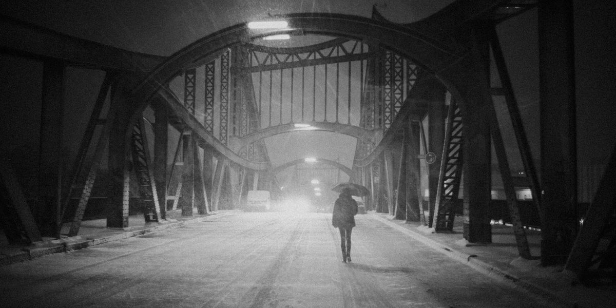 In a black and white photo a woman walks alone on a desolate bridge, in the snow, holding an umbrella