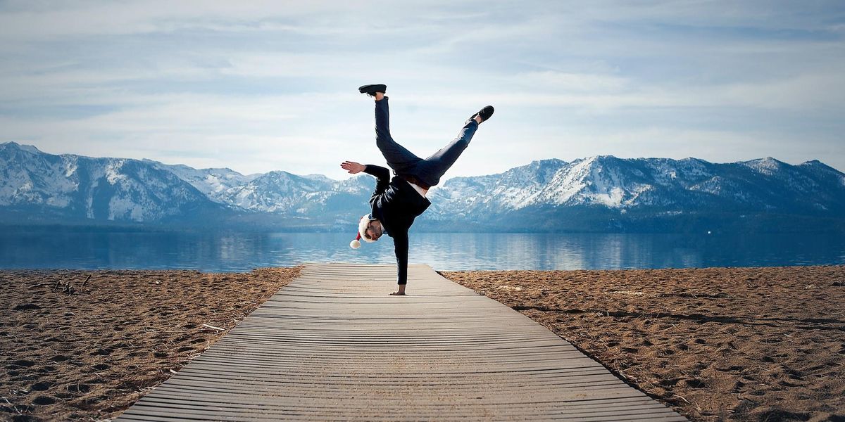 A guy in a Santa hat does a one armed hand stand with a mountain view behind him