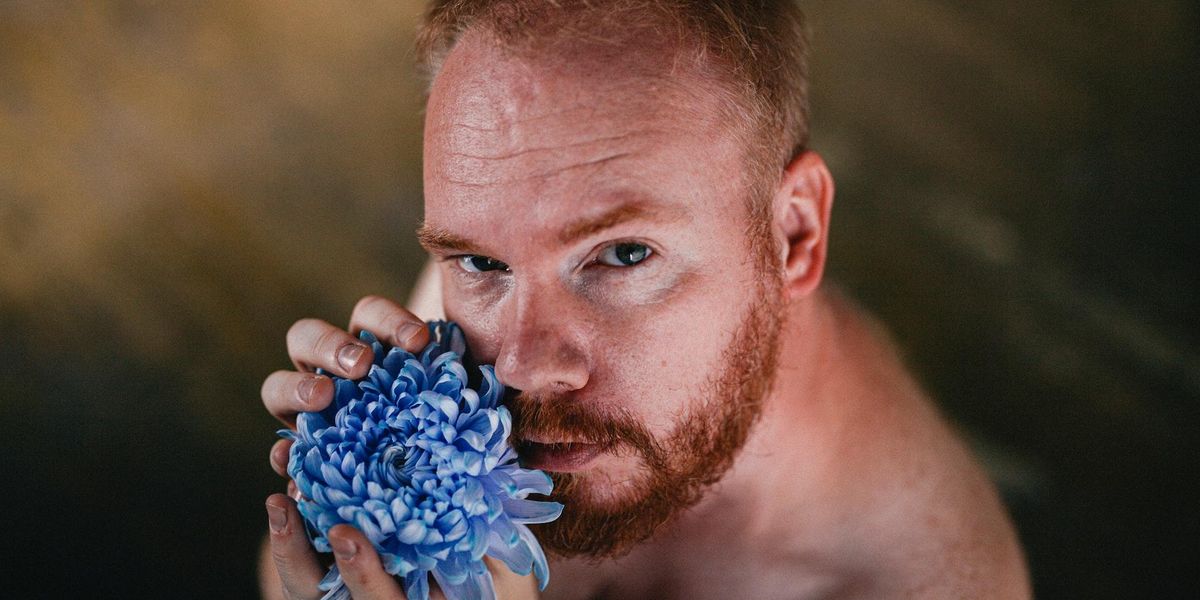 A red headed man holds a blue flower and seductively looks into the camera