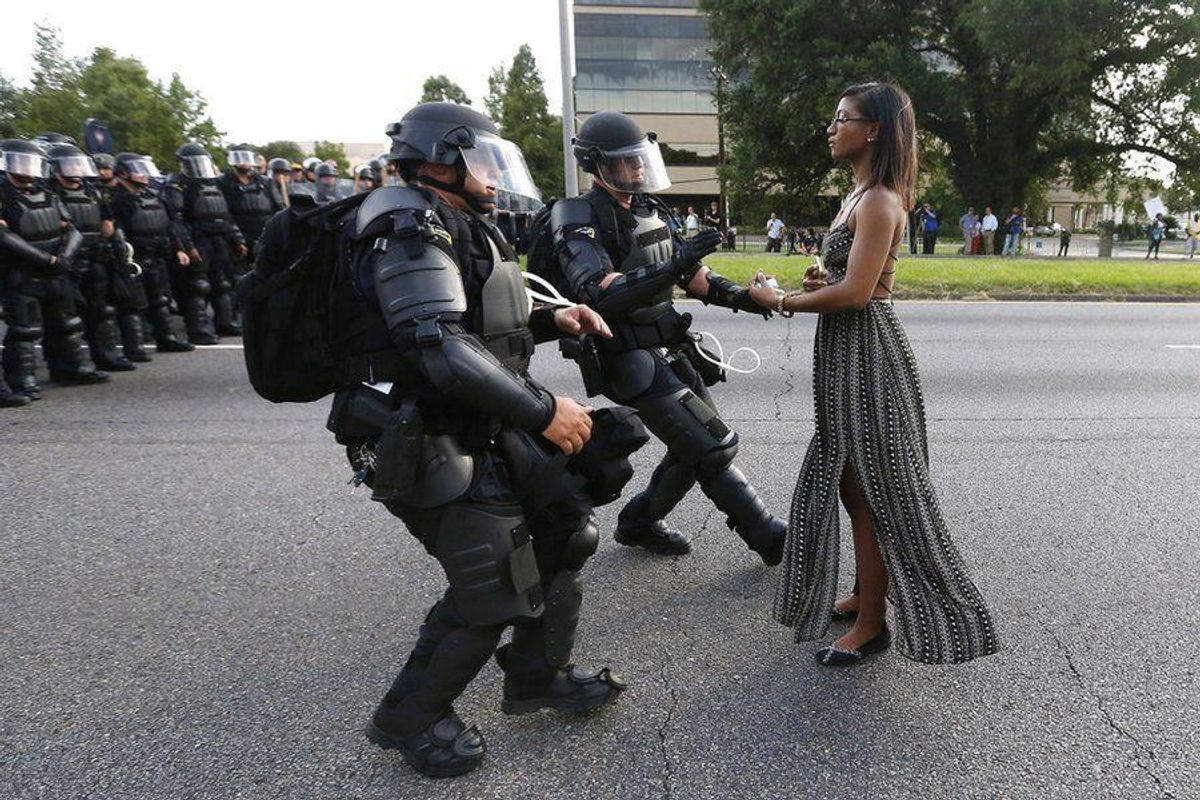 a woman stands calmly before two police officers in riot gear.