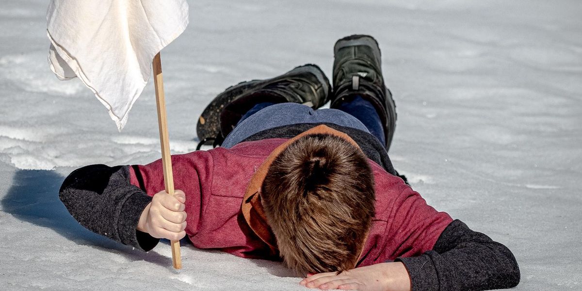 A young man lays face down on ice holding a white flag