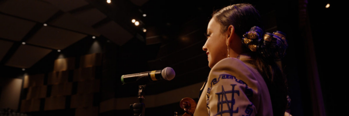 woman wearing a mariachi suit stands in front of a microphone facing an audience