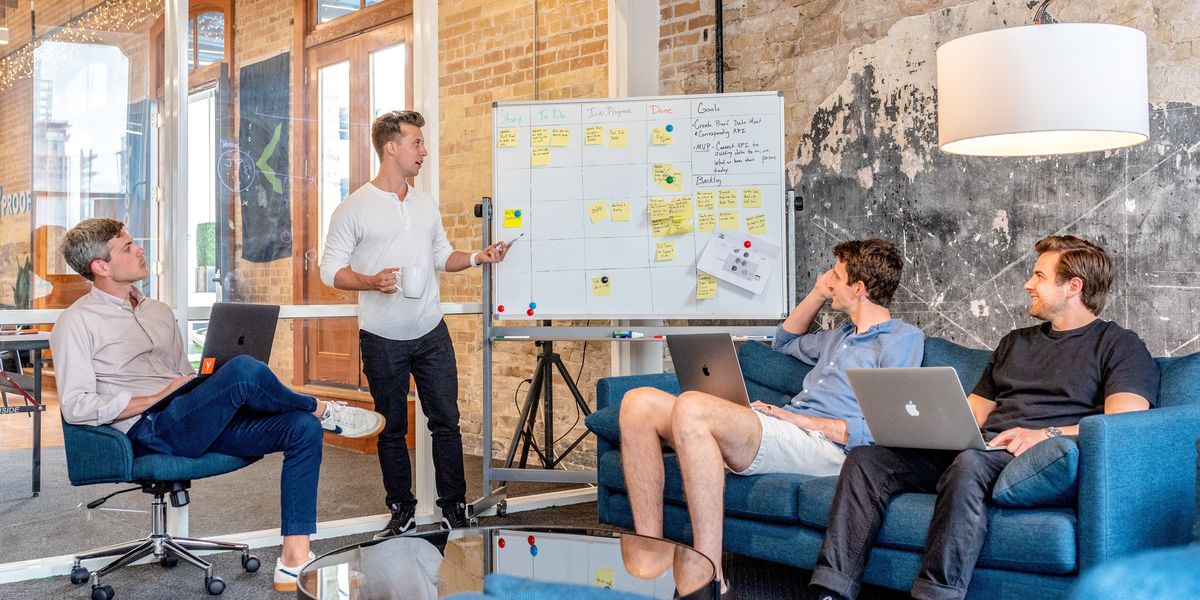 three men sitting while using laptops and watching man beside whiteboard