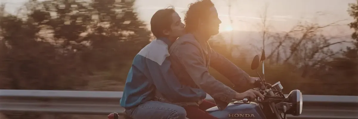 Still image from the film Too Late to Die Young / Tarde Para Morir Joven (2018) directed by Dominga Sotomayor showing a woman hugging a man from behind while riding a motorcycle with the sun behind them