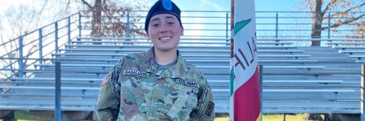 female solder in green fatigues stands in front of bleachers facing camera while smiling