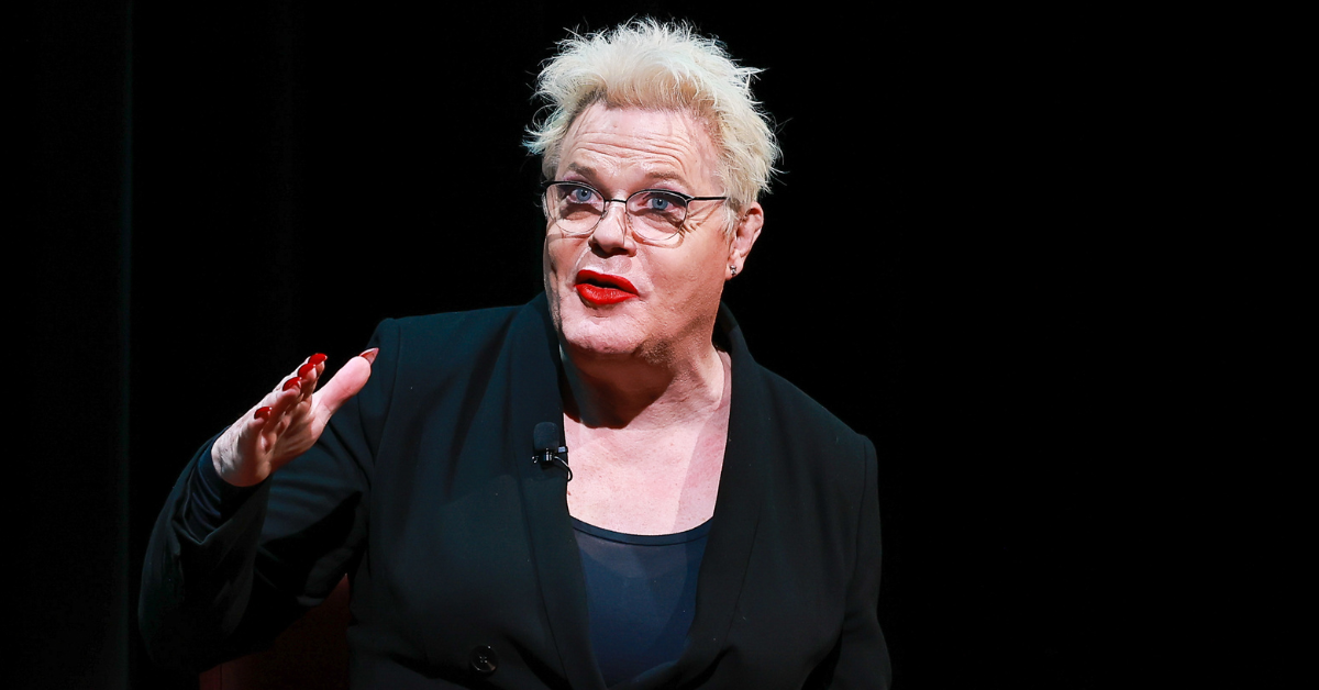 Comedian Suzy Eddie Izzard sits on a stage in front of a black background. She is in the middle of speaking and is gesturing with her right hand.