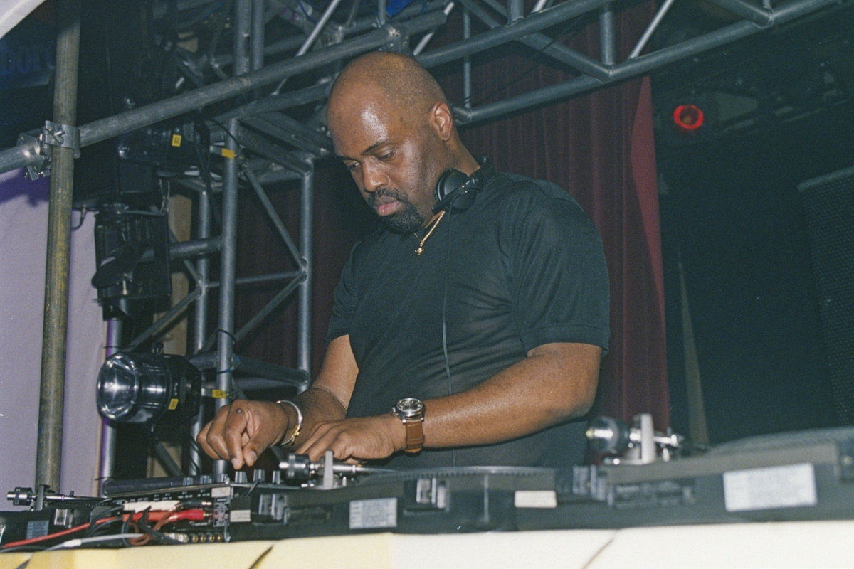 A DJ performing at a live event, focused on mixing tracks on a turntable, with a lit stage backdrop.