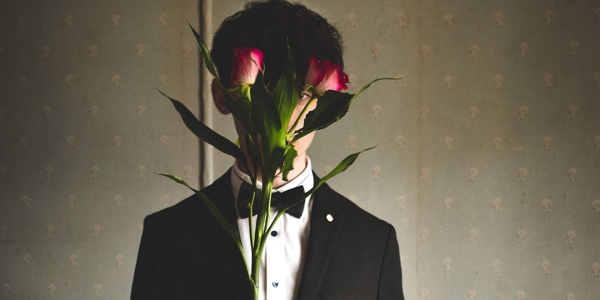 A young man in a tux, oddly holds two red roses in front of his face, only exposing one eye