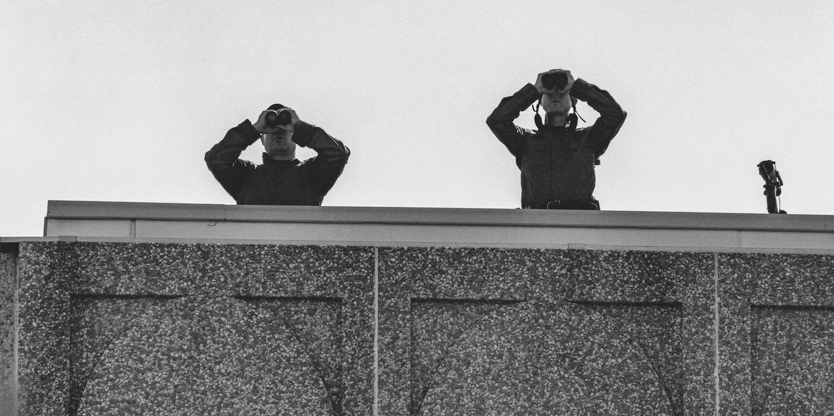 Two security people look out with binoculars from a roof