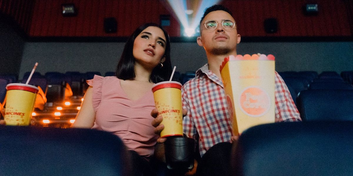 ​A couple watch a movie, in a theater, holding popcorn and two drinks