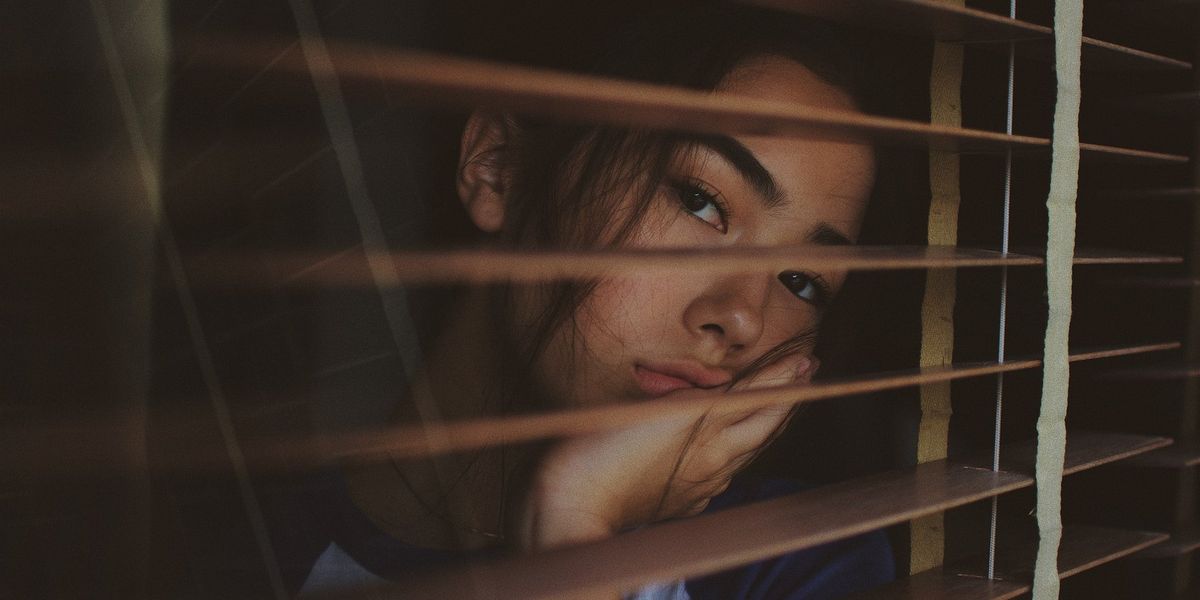 A young woman looks longingly out a window, through blinds