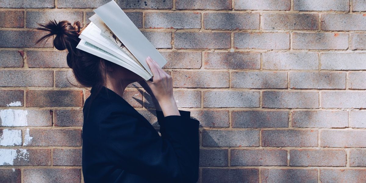 A seemingly frustrated woman stands in front of a brick wall, covers her face in book