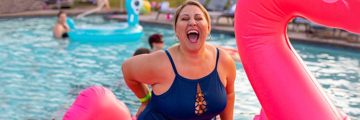 woman in a blue swimsuit sitting on a pink flamingo inflatable in a pool and laughing out loud