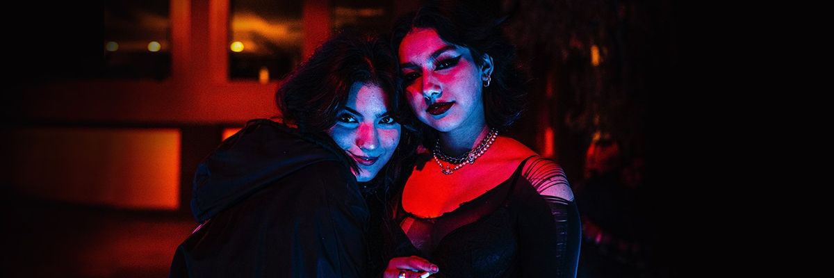 two goth girls in dark clothing and makeup smiling in moody lighting