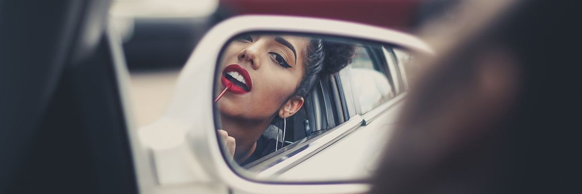 woman with big hoop earrings applying red lipstick on a car rearview mirror