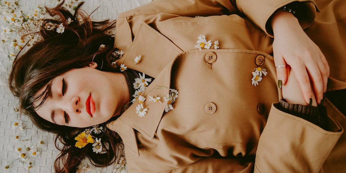 A woman lies on the ground, resting peacefully in a brown suede jacket, covered in small daisies