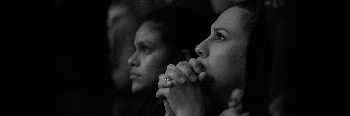 a woman clutching her hands together and looking up a something. another woman is in the background looking up.