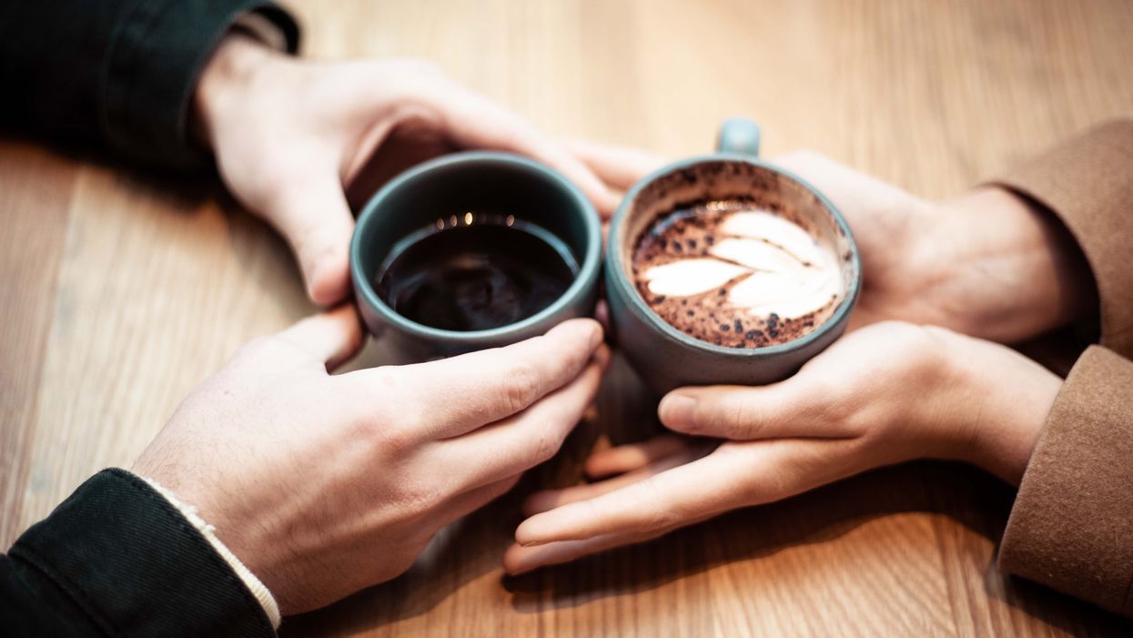 two people's hands holding ceramic mugs with coffee on wooden table