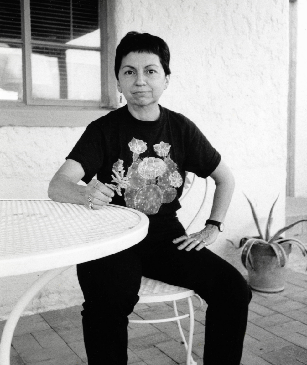 a black and white image of Gloria Anzald\u00faa sitting on a patio chair and table