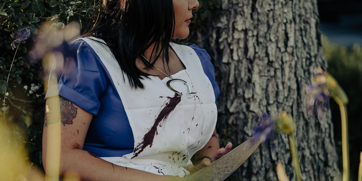 A woman hides behind a tree while she holds a knife and her front in splattered in blood