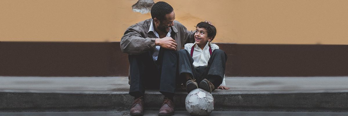 A father and his son sitting on a sidewalk smiling to each other while the boy has a soccer ball under his feet