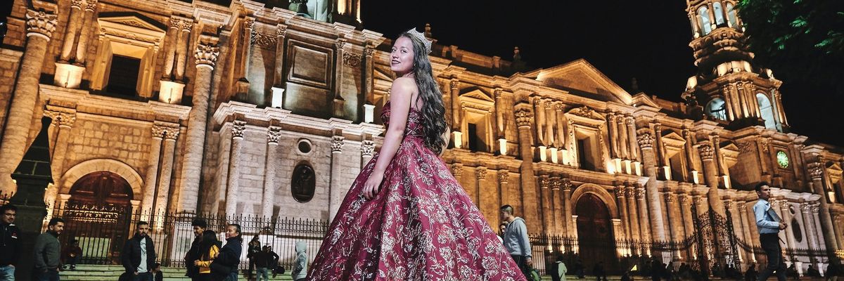 A quincea\u00f1era in a burgundy and gold gown posing in front of a building