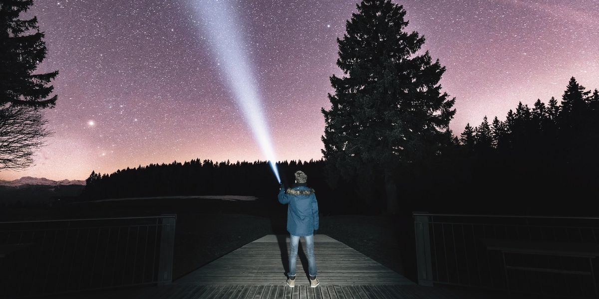 A guy stands on a dock, he holds up a flashlight into the night sky, stargazing