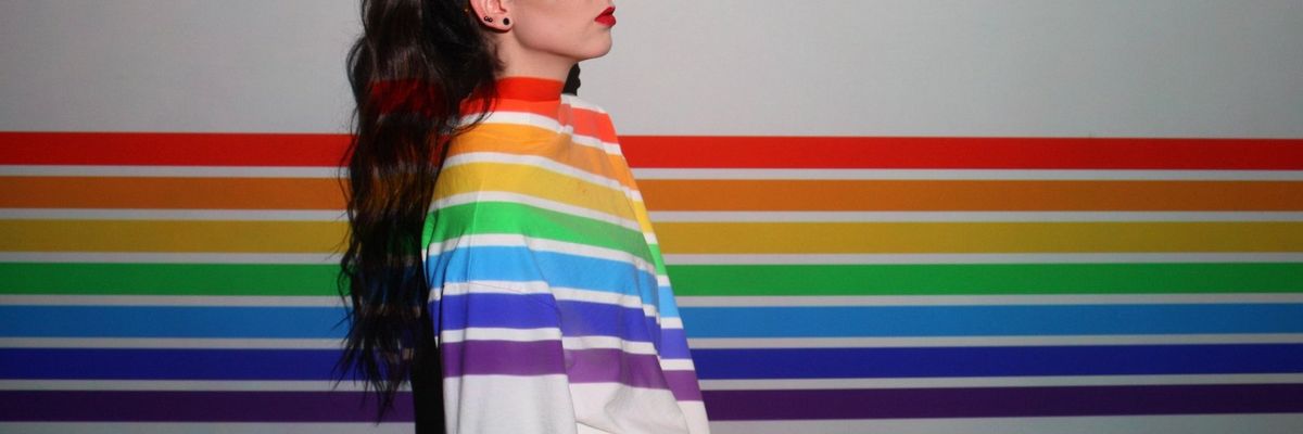A woman wearing orange glasses stands against a white wall with the pride rainbow painted on it and her white jacket