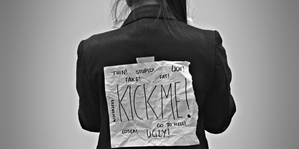 Black and white photo of a girl with a kick me sign, littered with expletives taped to her back