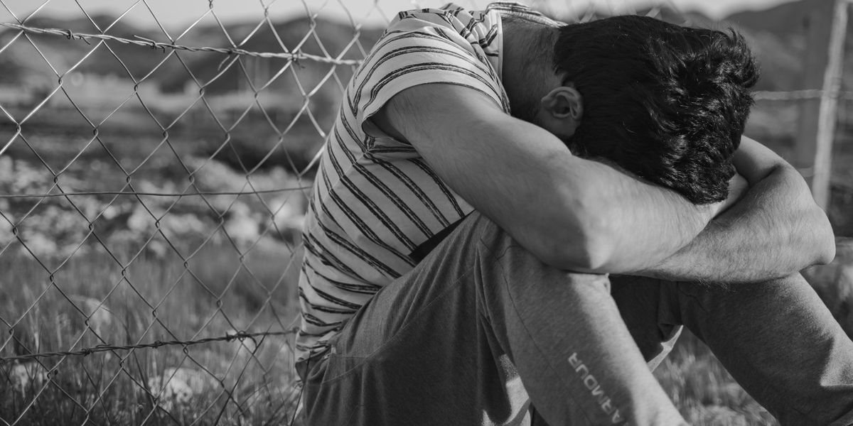 Man in a black and white stripe t-shirt sits along a chain link fence, with his head in his knees