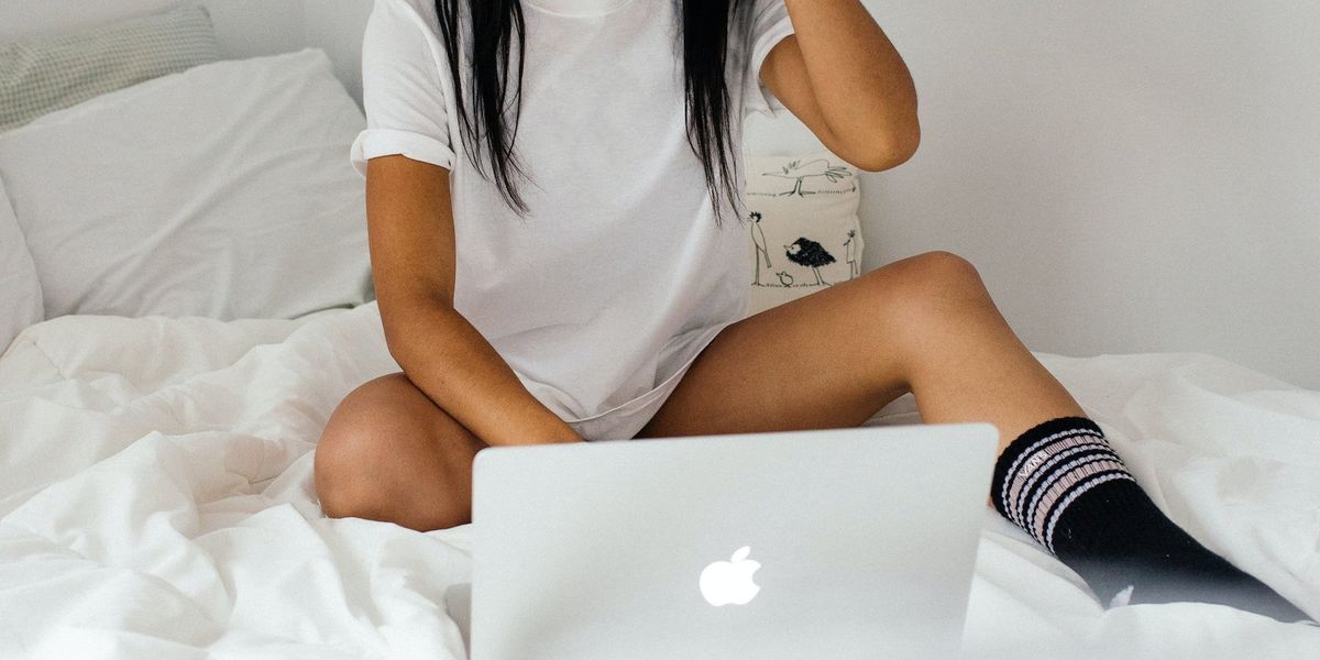 A young woman sits on a bed scrolling through a computer