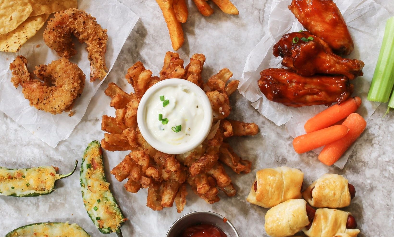 A wide variety of tailgate snacks are displayed on a counter top: Jalapeño poppers, fried onion bloom, pigs in a blanket, buffalo chicken wings, fried shrimp and fries.